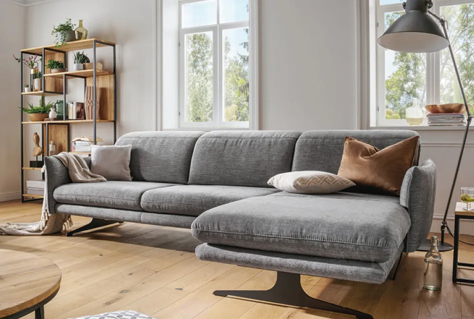 Graues Ecksofa mit Ottomane im modernen Wohnzimmer mit großen Fenstern, Holzregal und dekorativen Kissen. Beispiel für stilvolle Wohnlandschaft und komfortable Wohnzimmermöbel